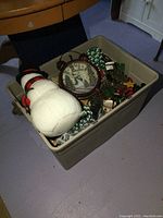 Photo showing a beige plastic bin filled with Christmas decorations including a large white plush snowman, a brown vintage-style alarm clock with Christmas snowman face, green and white ceramic trees, and assorted other items.