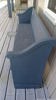 Side angled view of blue painted wooden church pew bench on a porch, showing its length and curved armrest design.