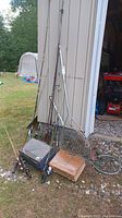 Photo showing multiple fishing poles leaning against a metal shed, two tackle boxes on the ground, and fishing nets on the side.