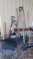 Photo showing assorted shovels, rakes, and sprayer leaning against a wall on gravel. Snow shovels, spades and rakes are visible.
