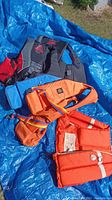 Four life jackets laid on blue tarp showing two Fluid brand jackets, one Buoy-O-Boy orange jacket, and one orange jacket with label.