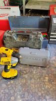 Yellow DeWalt cordless drill on table in front of gray Dremel case and blue metal toolbox
