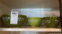 Three green Pyrex bowls of different sizes lined up on a shelf, showing their white floral pattern.