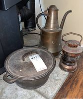 Cast iron Dutch oven, metal pitcher, and metal lantern grouped together on a surface, showing wear and rust.