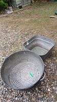Two vintage galvanized metal wash tubs placed on gravel outside. One round tub in the front and one square tub behind it.