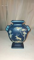 Front side of vase showing hand painted deer and floral scene in cream and yellow tones on a matte dark blue background.