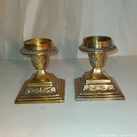 Pair of ornate brass candleholders with square bases and decorative swag ribbon patterns.