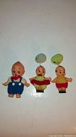 Three vintage Kewpie dolls with knit clothing and crochet hats, displaying different colors and sizes, on a white surface.
