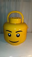 Front view showing yellow LEGO minifigure head container with handle and face print.