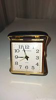 Front-facing view of the Phinney Walker compact travel alarm clock, displaying white clock face with black numerals and three hands, inside a gold-tone metal frame with black casing.