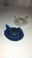 Pair of glass hen-on-nest salt holders, one cobalt blue and one clear glass, placed on flat surface.