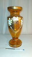 Front view of large amber glass vase showing raised white flowers and gold design with pedestal base, pen placed for size reference.