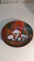 Top view of round art plate with mushroom design in shades of white, yellow, orange, red, green, and blue on copper base.