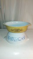 Two vintage Pyrex bowls stacked, showing the yellow mixing bowl with brown and orange pattern on top and the white casserole with blue floral pattern underneath