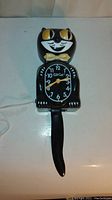 Front view of the Kitcat clock showing black cat body, yellow eyes and bow tie, clock face with white numbering and yellow hands, tail pendulum, plugged in cord.