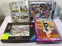 Four boxed jigsaw puzzles displayed showing front covers: Alan Giana set, world landmarks collage 1000-piece, Ravensburger winter village 1000-piece, and Naruto Morita Geisha 1000-piece.
