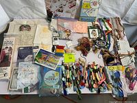 Overview photo showing wide assortment of cross-stitch supplies including multiple embroidery floss skeins, patterns, and fabric pieces arranged on a white surface.