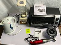 Photo showing the assembled set of kitchen appliances on a white surface: Black & Decker toaster oven, Oster blender, white electric kettle, red immersion blender, and a chopper.