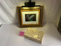 Framed Celebrate print with ornate gold frame and LED battery powered picture light illuminated above. Vintage sheet music covered wood box in front.