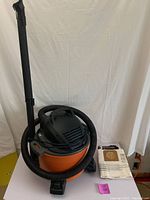 Orange and black Ridgid 6 gallon shop vacuum with hose and floor wand attached, against white backdrop.