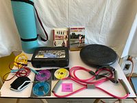 Overview photo of the exercise equipment lot on a white table against a white backdrop. Includes blue rolled exercise mat, two exercise books, three DVDs with colorful labels, two resistance bands with gray foam handles, jump ropes, black air-filled balance disk, and small black exercise glove.