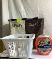 Overall laundry lot showing divided hamper with 'Lights' and 'Darks' sections, white laundry basket, and tub of Tide Power Pods