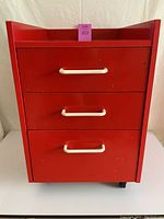 Front view of vintage red lacquer rolling drawer unit with three drawers and white handles showing some wear and chips.