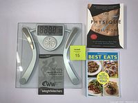 WeightWatchers WW71C electronic body fat scale on glass platform with digital display and 5 control buttons. Next to scale are two paperback books: 'The Physique Solution' fitness book and 'WeightWatchers Best Eats' cookbook.