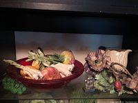 Red bowl holding assorted porcelain artificial fruit and vegetables including leeks, apples or onions, and carrots with a cluster of green grapes next to the bowl.