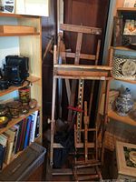 Image showing multiple artist easels of varied sizes and types stored upright against a wall, including wooden and metal components.