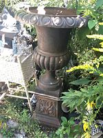 Full front view of tall Grecian urn on square scroll-relief base surrounded by garden foliage