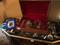 Wide shot showing the closed wood flatware case with silver cutlery inside and loose utensils to the right, including a whiskey flask.