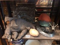 Overall lot view on wooden shelf showing dragon figurine, ceramic egg, metal plaque and snail lamp