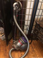 Dyson vacuum cleaner upright with hose coiled, placed on hardwood floor next to cabinet and metal fence structure.