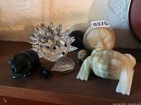 Collection of animal figurines including clear crystal spiked hedgehog, black stone frog with lily pad, beige stone frog, and other small carved stone animals on wooden surface.