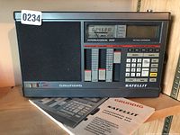 Front view of Grundig International 400 short wave radio showing speaker, digital tuning display, sliders and control buttons, with paperwork/manuals underneath.
