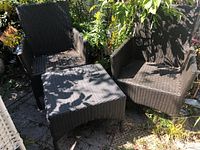 Photo of two dark brown woven patio chairs and matching square woven table on outdoor stone patio with greenery in background