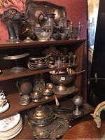 Shelf with assorted silver plated trays, bowls, pitchers, serving pieces, and decorative items showing tarnishing and patina.