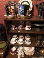 Shelving unit with stacked decorative plates, two boxed mugs, and multiple sets of teacups and saucers arranged on shelves.