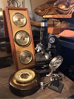 Photo showing vintage black microscope, wooden plaque with three instruments, round wooden base barometer, and magnifying stand.