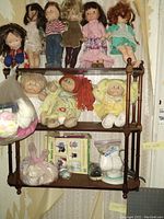 Full view of dolls on wooden three-tiered wall shelf showing cloth dolls, Cabbage Patch dolls, paper doll set and accessories