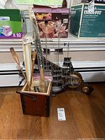 View of wooden box with various wooden kitchen tools inside along with a wooden ship model and wooden clock behind on floor