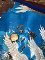 Close-up photo of a vintage square-faced wristwatch casing, gold-tone ring, cufflink, and rectangular pin on blue bird-patterned shawl.