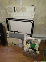 Photo of white foldable Americrafters craft table with black legs, plastic organizer tray, box of art supplies and paper products leaning against a wall.