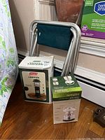 Boxed Coleman propane lantern, boxed Northpoint multifunction radio lantern, and foldable stool with green fabric seat leaning against wall