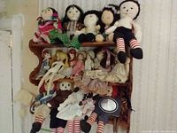 Photo showing a collection of fabric rag dolls seated and arranged on a wooden shelf, including vintage Raggedy Ann and Holly Hobbie dolls.