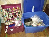 Wide shot showing wooden thread rack loaded with various spools of thread, maroon wicker sewing box next to it, and blue plastic bin filled with assorted sewing accessories on wooden floor.