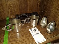 Two silver plated creamers or cups with handles, a pair of salt and pepper shakers, and a dark fabric pouch placed on a wooden surface.