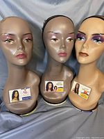 Front view of three long neck wig heads with detailed painted facial features and black wig nets. Each head has a different makeup style and skin tone.