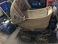 Side view of antique wicker baby pram showing woven wicker body, glass side window, metal frame, large spoked wheels, and interior fabric lining.